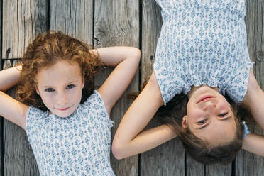 From above shot of two sweet sisters in similar dresses smiling and looking at camera while lying on lumber surface on sunny day - ADSF40424