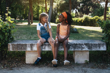 African American girl smiling and best friend sitting together on bench in green park in sunny day - ADSF40432