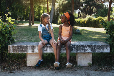 Afroamerikanisches Mädchen lächelnd und beste Freundin sitzen zusammen auf Bank in grünen Park in sonnigen Tag - ADSF40432