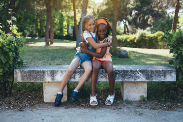 African American girl smiling and embracing to best friend sitting together on bench in green park in sunny day - ADSF40433