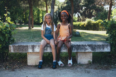 African American girl smiling and holding hand with best friend sitting together on bench in green park in sunny day - ADSF40434