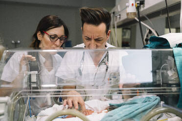 Ein Ärztepaar kümmert sich aufmerksam um ein gesichtsloses neugeborenes Baby, das in einem Inkubator in einer Klinik liegt - ADSF40455