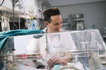 Männlicher Arzt kümmert sich aufmerksam um ein gesichtsloses neugeborenes Baby, das in einem Inkubator in einer Klinik liegt - ADSF40459