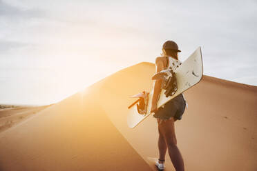 Rückenansicht einer jungen Frau, die auf Sand läuft und sich auf das Sandboarding vorbereitet und dabei wegschaut - ADSF40486