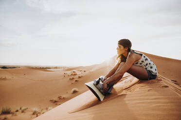 Seitenansicht einer jungen Frau mit Mütze und Sonnenbrille, die auf Sand sitzt und sich auf das Sandboarding vorbereitet - ADSF40488