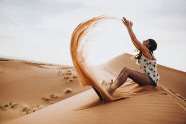 Seitenansicht einer jungen Frau mit Mütze und Sonnenbrille, die auf Sand sitzt, diesen in die Luft wirft und sich auf das Sandboarding vorbereitet - ADSF40489