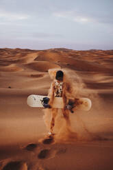 Rückenansicht einer nicht erkennbaren jungen Frau, die auf Sand steht und sich auf das Sandboarding vorbereitet - ADSF40490