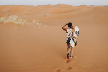 Rückenansicht einer jungen Frau, die auf Sand läuft und sich auf das Sandboarding vorbereitet und dabei wegschaut - ADSF40493