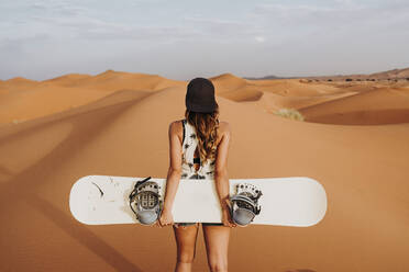 Rückenansicht einer jungen Frau, die auf Sand läuft und sich auf das Sandboarding vorbereitet und dabei wegschaut - ADSF40494