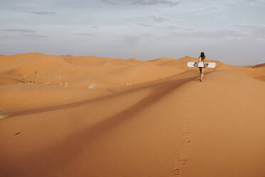 Rückenansicht einer jungen Frau, die auf Sand läuft und sich auf das Sandboarding vorbereitet und dabei wegschaut - ADSF40495