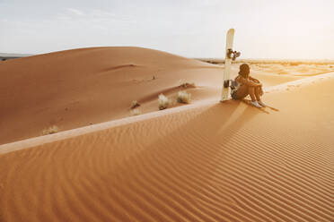 Seitenansicht einer nicht erkennbaren jungen Frau mit Mütze, die auf Sand sitzt und die Wüstenlandschaft beim Sandboarding bewundert - ADSF40497