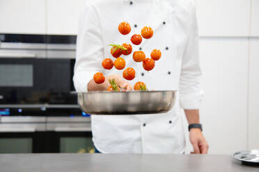 Ein Mann in weißer Kochjacke hält eine Pfanne und mischt Kirschtomaten mit Kräutern, während er in einer Restaurantküche steht - ADSF40498