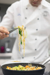 Crop anonymer Koch in weißer Uniform Kochen von Nudeln mit Gemüse in der Pfanne auf dem Herd auf dem Tisch in der hellen Küche - ADSF40500