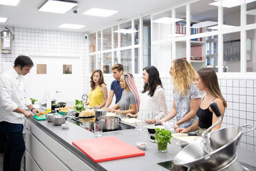 Gruppe multiethnischer Menschen, die am Tisch stehen und lernen, während ein Mann den Prozess des Gemüseschneidens in einer professionellen Küche demonstriert - ADSF40507