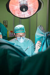 Männlicher Arzt mit Brille und Uniform, der mit Hilfe von Assistenten in einem modernen Operationssaal eines Krankenhauses einen Patienten operiert - ADSF40517