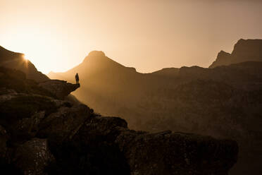 Back view of unrecognizable female traveler silhouette sitting on edge of cliff and admiring picturesque mountains in morning in Pyrenees - ADSF40531