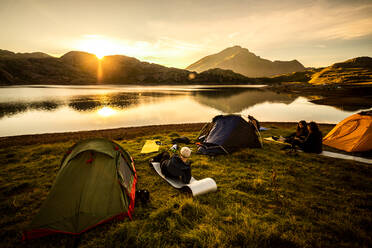 Group of traveling friends with camping tents sitting on grassy shore near calm lake reflecting mountains and enjoying spectacular sundown in summer evening - ADSF40549