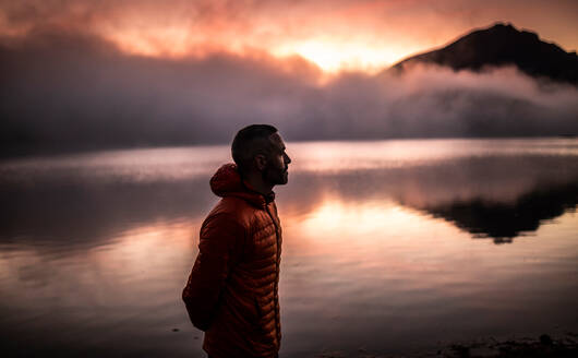 Side view of lonely man in outerwear looking away thoughtfully while relaxing near calm lake water reflecting mountains and cloudy sky at sunset time - ADSF40550