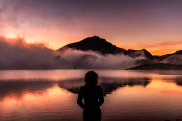 Back view of lonely unrecognizable woman in outerwear looking away thoughtfully while relaxing near calm lake water reflecting mountains and cloudy sky at sunset time - ADSF40551