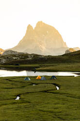 Eine Gruppe von Reisefreunden mit Campingzelten sitzt am grasbewachsenen Ufer in der Nähe eines ruhigen Sees, in dem sich die Berge spiegeln, und genießt den spektakulären Sonnenuntergang am Sommerabend - ADSF40552