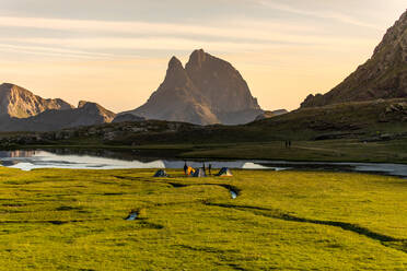 Group of traveling friends with camping tents sitting on grassy shore near calm lake reflecting mountains and enjoying spectacular sundown in summer evening - ADSF40553