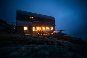 Eine Gruppe unerkennbarer Abenteurer sitzt auf der Terrasse eines Gästehauses mit beleuchteten Fenstern in einer nebligen Nacht in einer Bergregion - ADSF40558