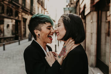 Side view of joyful lesbian couple with piercing and tattoos kissing on street - ADSF40567