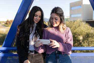 Female friends standing on bridge and surfing Internet on smartphone while enjoying weekend in city on sunny day - ADSF40584