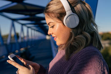 Nahaufnahme einer fröhlichen Frau mit Kopfhörern, die auf einer Brücke steht, während sie Musik hört und mit ihrem Smartphone im Internet surft - ADSF40586