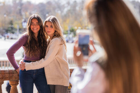 Frau fotografiert Frauen, die sich umarmen und auf der Plaza de Espana stehen, während sie ihr Smartphone benutzt - ADSF40592