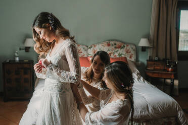 Attraktive glückliche Damen, die einer jungen Frau in der Nähe des Bettes im Schlafzimmer beim Anziehen des Hochzeitskleides helfen - ADSF40695