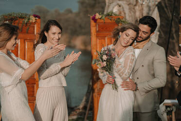 Junger Mann in grauem Kostüm umarmt attraktive lächelnde Dame in Hochzeitskleid und Blumenstrauß in der Nähe von Dekorationen und Freunde klatschen Hände in der Nähe von See - ADSF40702