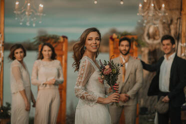 Porträt einer fröhlichen Braut mit Blumenstrauß bei einer Hochzeitsfeier, im Hintergrund feiernde Gäste - ADSF40705