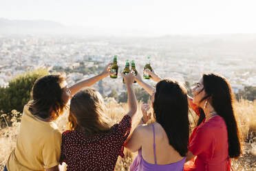 Back view of group of joyful female friends with arms raised toasting and clinking glass bottles of beer while sitting on hilltop over town against cloudless sky in sunlight - ADSF40715
