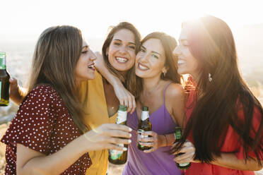 Delighted female friends in stylish outfits with bottles of alcohol beverages having fun while standing on hilltop over town against cloudless sky in sunlight - ADSF40717