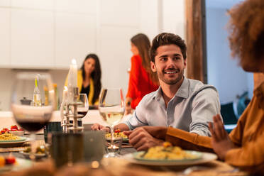 Affectionate smiling young man holding hand of girlfriend while sitting together at festive table with dishes and glasses of wine during home party with friends - ADSF40724