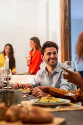 Affectionate smiling young man holding hand of girlfriend while sitting together at festive table with dishes and glasses of wine during home party with friends - ADSF40725
