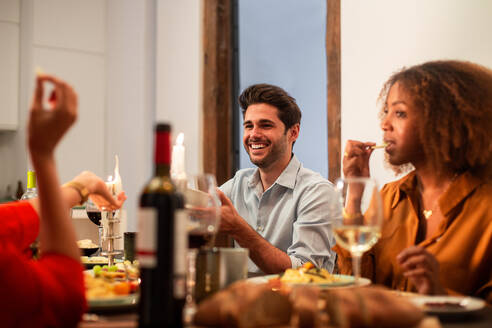 Eine Gruppe moderner junger multirassischer Freunde versammelt sich an einem festlichen Tisch mit köstlichen Gerichten und Wein und genießt die gemeinsame Zeit zu Hause - ADSF40726