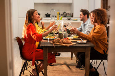 Side view of cheerful multiracial friends sitting at festive table and eating pasta during celebration and party in modern apartment - ADSF40727