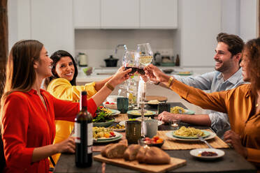 Seitenansicht eines fröhlichen, bunt gemischten Freundeskreises, der feiert und mit Weingläsern anstößt, während er an einem gedeckten Tisch mit einem köstlichen Abendessen zu Hause sitzt - ADSF40728