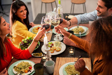 Von oben Seitenansicht von fröhlichen, verschiedenen Freunden, die feiern und mit Weingläsern anstoßen, während sie an einem gedeckten Tisch mit einem köstlichen Abendessen zu Hause sitzen - ADSF40729