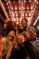 Young content diverse partners making faces while taking self portrait on cellphone under shiny lights during night celebration - ADSF40740