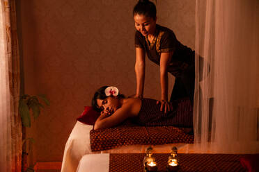 Asian female massagist in traditional clothes giving a thai massage to a young attractive woman with orchid on head lying on a bed stretcher in a room lit with warm lights - ADSF40751