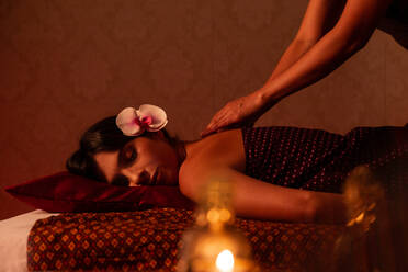 Crop view of asian female massagist giving a thai massage to a young attractive woman with orchid on head lying on a bed stretcher in a room lit with warm lights - ADSF40752