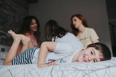 Von unten junger lachender Junge im Schlafanzug, der mit Kindern und Familie spielt und in die Kamera schaut, auf dem Bett im Schlafzimmer - ADSF40759