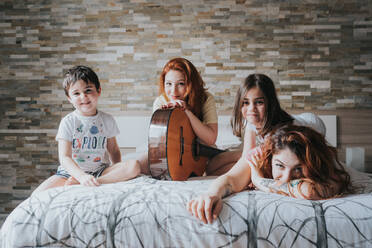 Junge gleichgeschlechtliche Eltern im Schlafanzug spielen Gitarre mit Kindern und schauen in die Kamera auf dem Bett im Schlafzimmer - ADSF40765