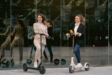 Zufriedene Unternehmerinnen in eleganter Freizeitkleidung stehen mit eklektischen Motorrollern in der Nähe eines Glasgebäudes in der Innenstadt und schauen weg - ADSF40775