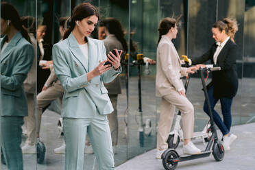 Determined female entrepreneur in suit standing near glass building and using smartphone on background of colleagues on eclectic scooters - ADSF40776