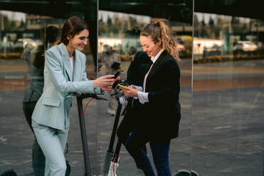 Cheerful female entrepreneurs standing near electric scooters in city and browsing mobile phones - ADSF40777