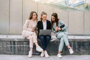 Gesellschaft von lächelnden Geschäftsfrauen in eleganter Freizeitkleidung, die in der Stadt sitzen und einen Laptop benutzen, während sie gemeinsam an einem Startup-Projekt arbeiten - ADSF40781